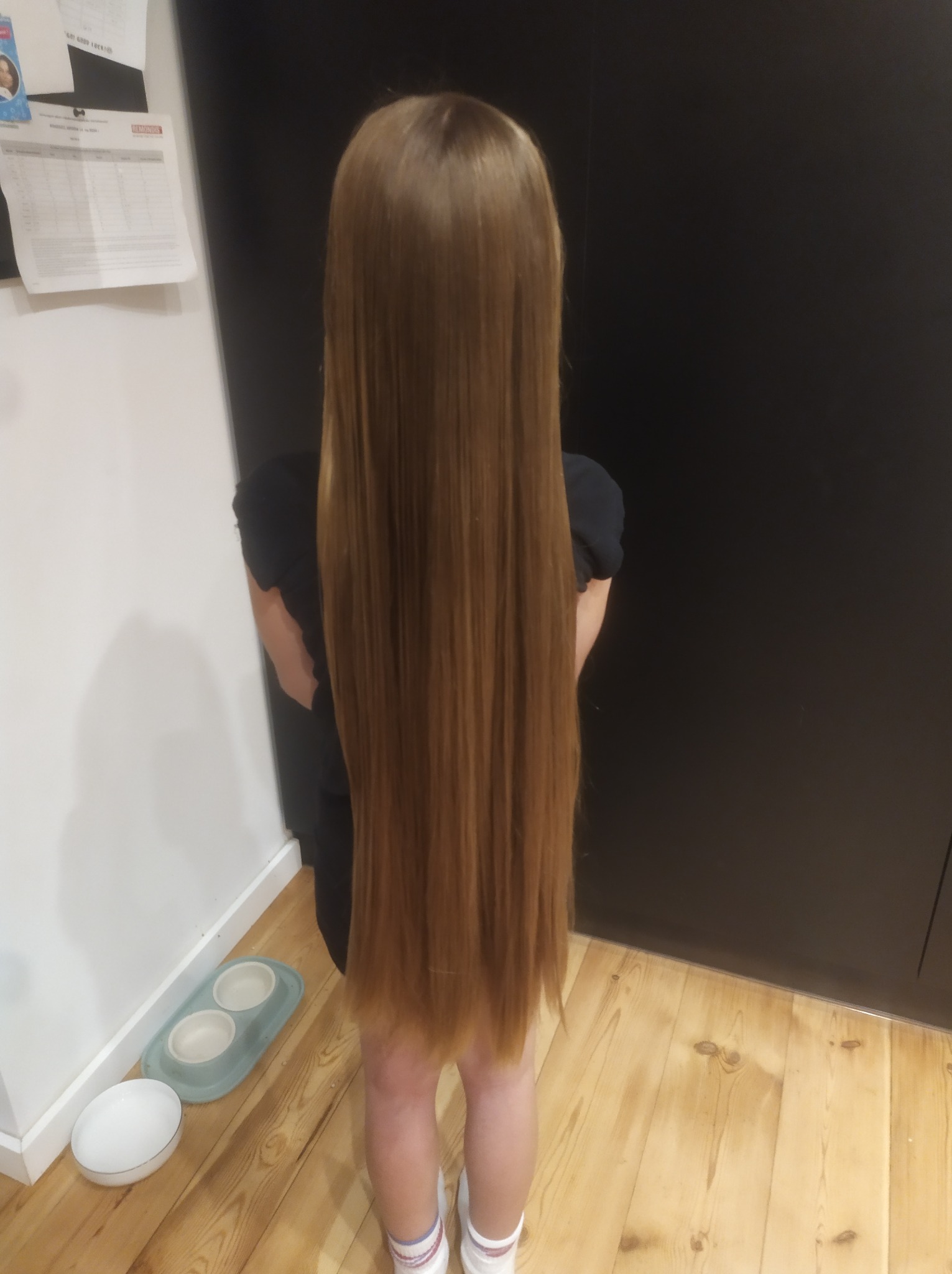 Dziewczynka z bardzo długimi, prostymi, brązowymi włosami, stojąca tyłem na tle drewnianej podłogi i ciemnej szafki.