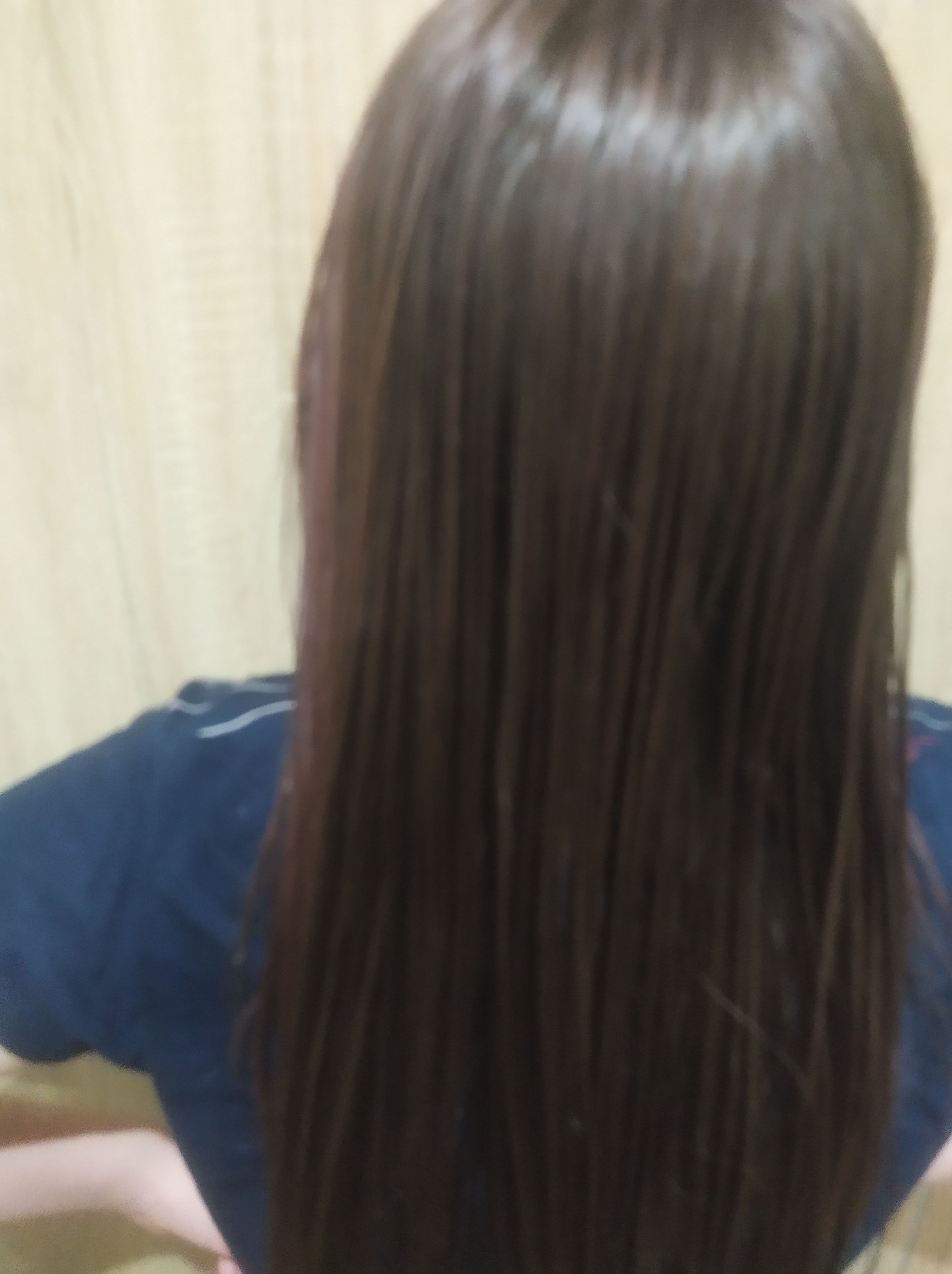 Widok z tyłu na długie, proste, brązowe włosy po zabiegu pielęgnacyjnym, z delikatnym połyskiem i minimalnym rozdwajaniem końcówek, na rozmytym tle ściany.