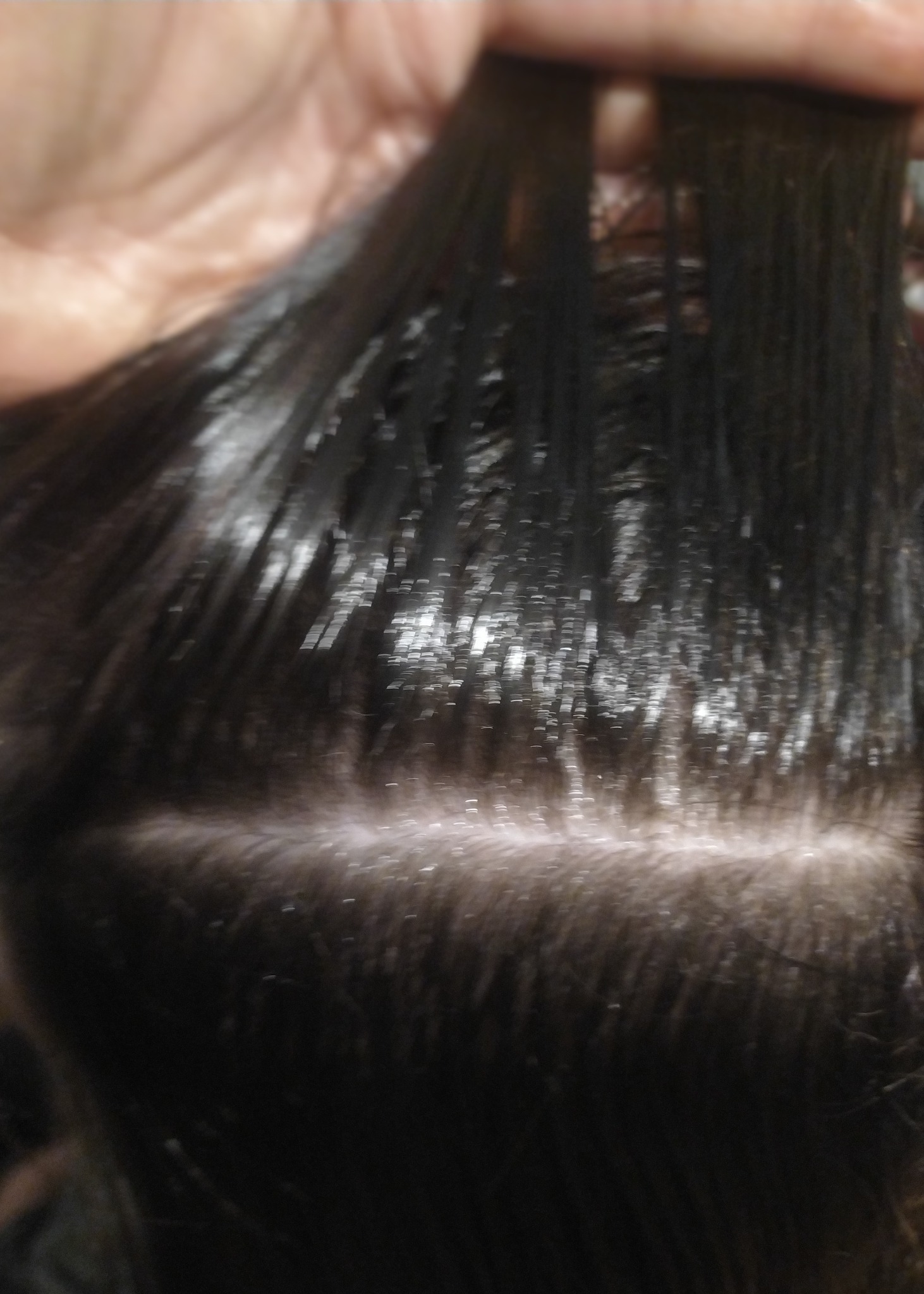 Proces nakładania preparatu na ciemne włosy przy przedziałku, widoczne pasma włosów pokryte błyszczącym produktem, trzymane w palcach.