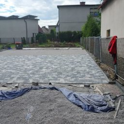 STAVO Katarzyna Kolon - Fragment nowej nawierzchni z szarej kostki brukowej ułożonej we wzór, z pracownikiem z wózkiem i narzędziami po lewej stronie, oraz stertą kostki w tle, widok na posesję z metalowym ogrodzeniem.