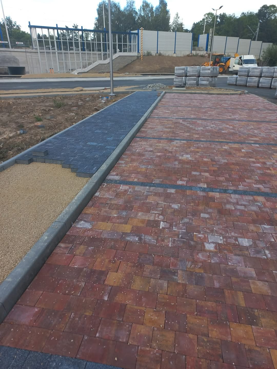 Częściowo ułożona nawierzchnia z kostki brukowej w odcieniach czerwieni i brązu, kontrastująca z fragmentem wykończonym ciemną kostką i podsypką piaskową, w tle widoczny wiadukt i maszyny budowlane.