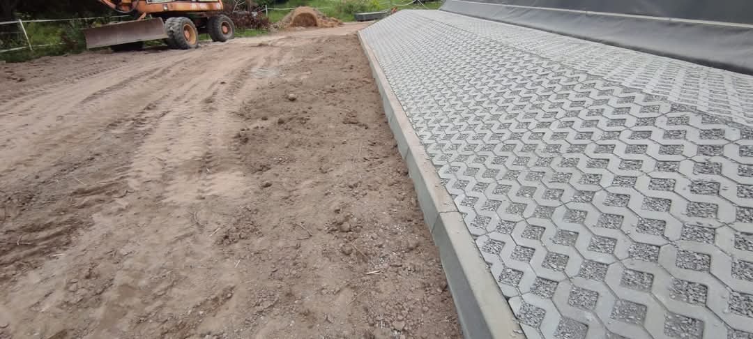 Układanie ażurowej kostki brukowej na podbudowie z kruszywa, widoczny fragment drogi gruntowej i ciężki sprzęt budowlany w tle.
