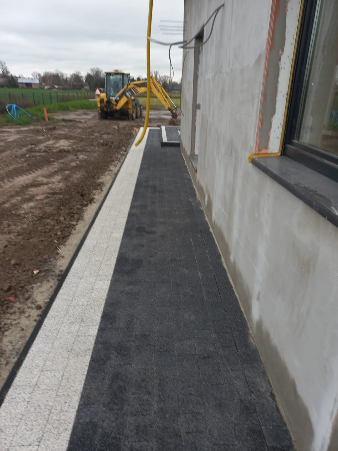 Nowo ułożony chodnik z kostki brukowej w kolorze czarnym i białym, przylegający do ściany budynku w trakcie wykończenia; w tle widoczna koparka na placu budowy.