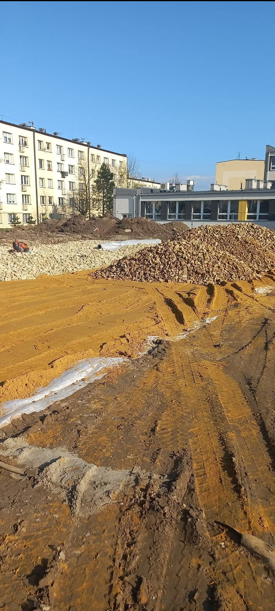 Teren budowy z widocznymi śladami opon, pryzmami kamieni i piasku, białą geowłókniną, budynkami w tle i błękitnym niebem.