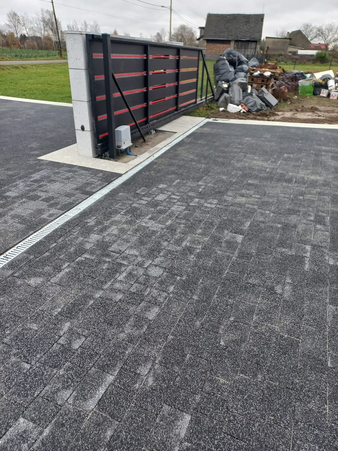 Ułożona szara kostka brukowa z odpływem liniowym, brama przesuwna z automatyką w tle, widoczne ślady wilgoci na kostce, w tle sterta śmieci i gruzu.