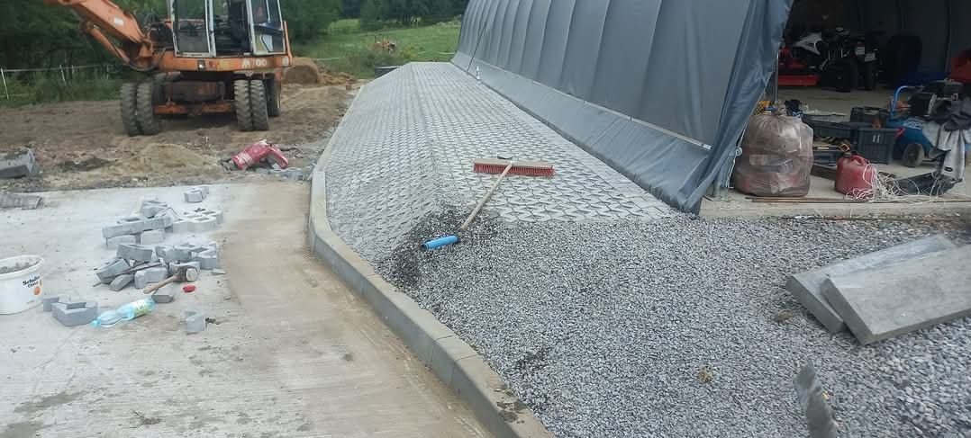 Układanie kostki na podjeździe z użyciem ażurowych płyt, widoczny rozsypany żwir, krawężnik, koparka w tle i akcesoria brukarskie.