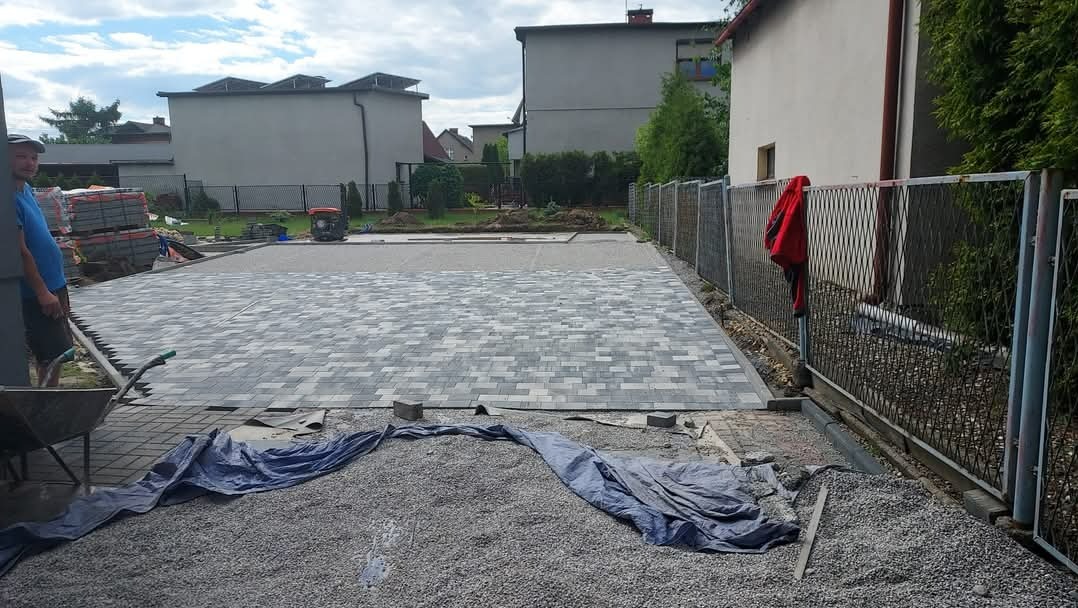 Fragment nowej nawierzchni z szarej kostki brukowej ułożonej we wzór, z pracownikiem z wózkiem i narzędziami po lewej stronie, oraz stertą kostki w tle, widok na posesję z metalowym ogrodzeniem.