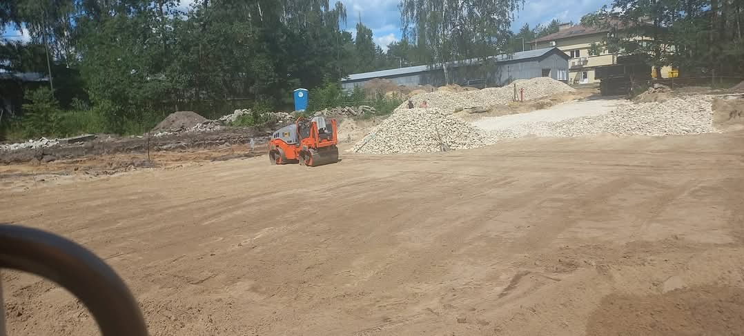 Pomarańczowy walec drogowy ugniata piasek na placu budowy, w tle hałda kamieni, widoczna toaleta przenośna i zabudowania.