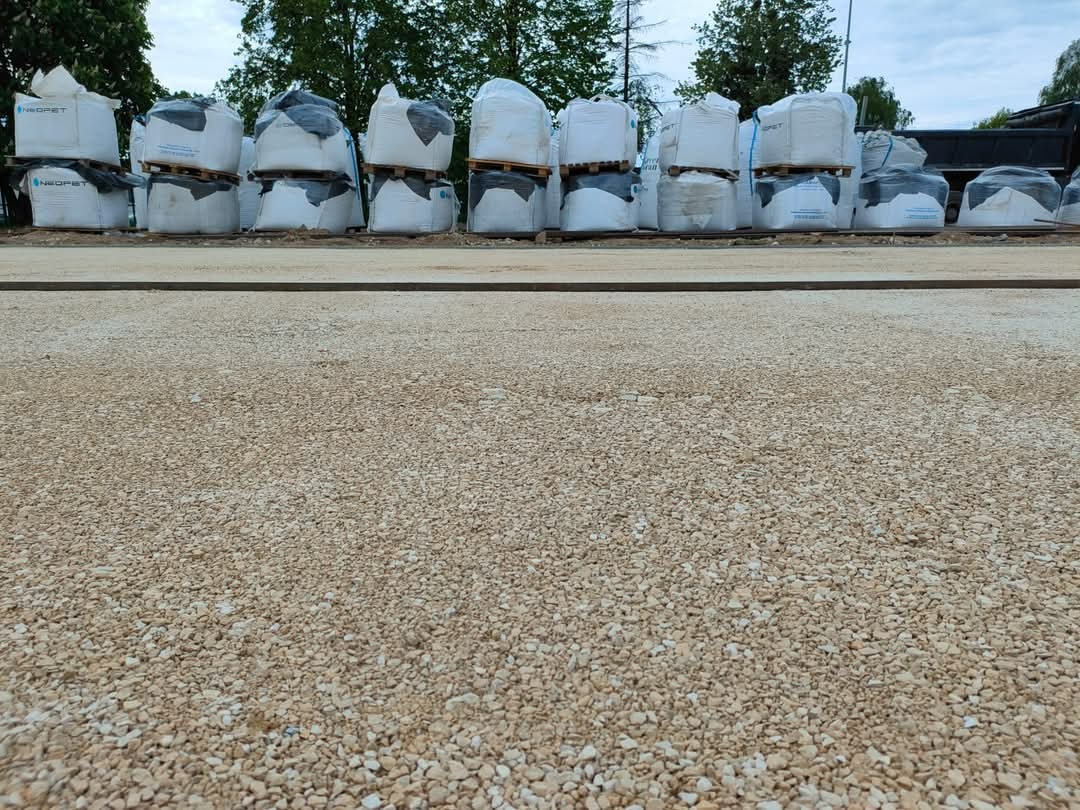 Przygotowany teren z drobnym kruszywem pod utwardzenie, w tle palety z workami materiału budowlanego, widoczne drzewa i fragment ciężarówki.