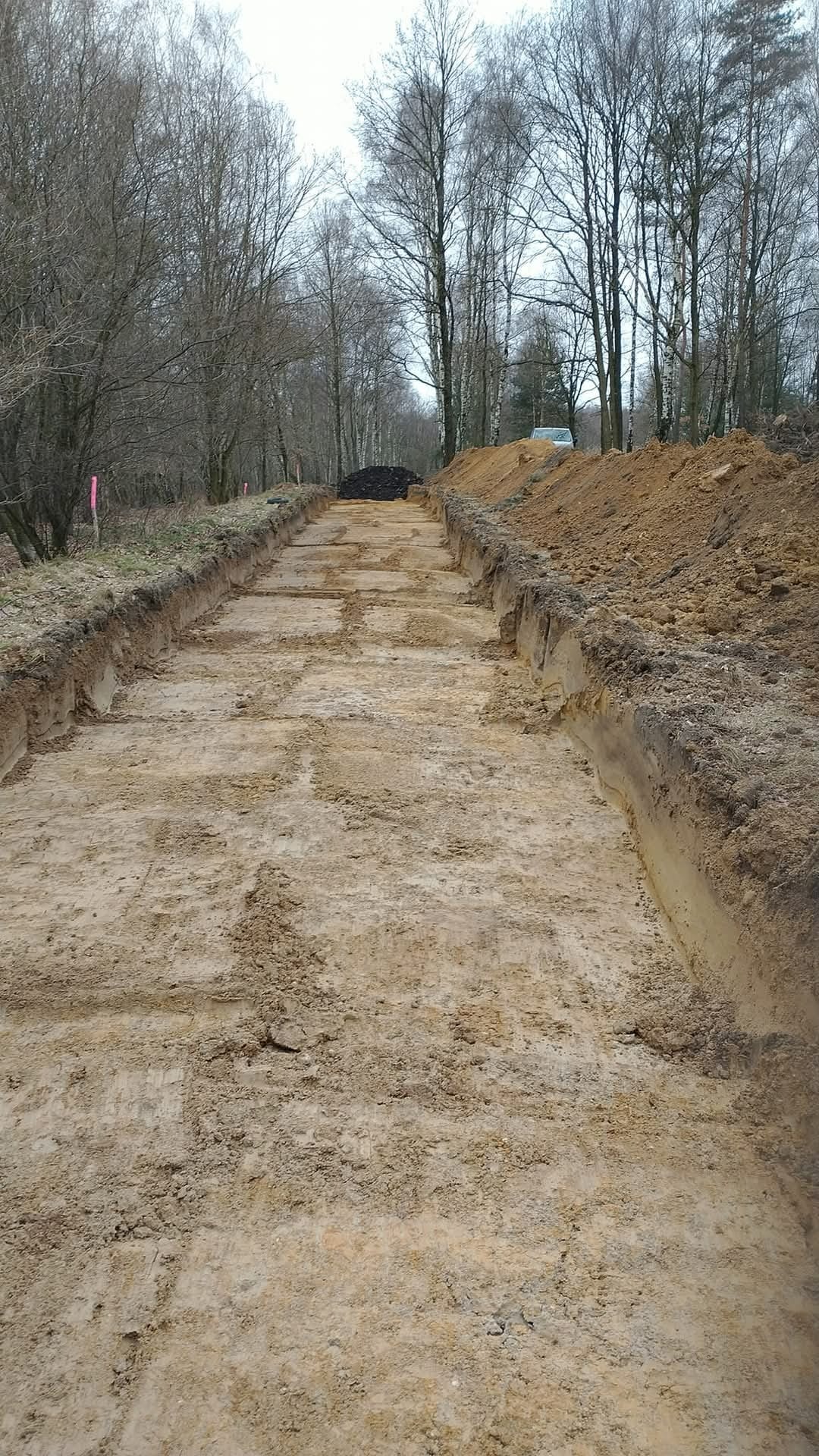 Wykopany rów w ziemi, widoczne ślady po maszynach, w tle drzewa i hałda ziemi.