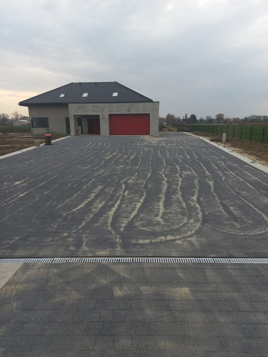 Nowo ułożona, szara kostka brukowa przed nowoczesnym domem z czerwoną bramą garażową; widoczny drenaż liniowy i pozostałości piasku na powierzchni kostki.