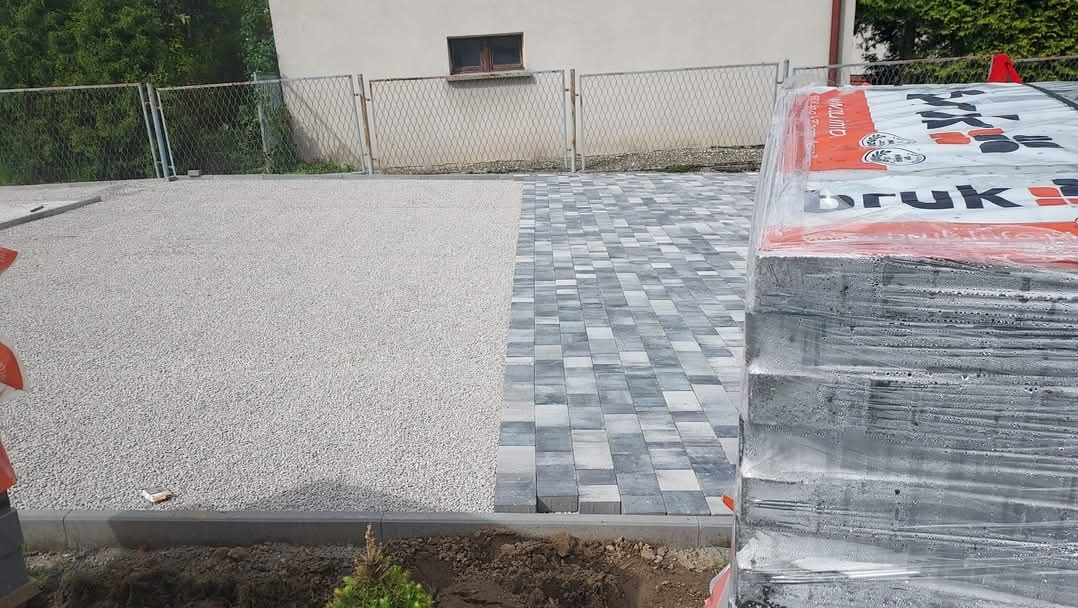 Częściowo ułożona szara kostka brukowa w geometryczny wzór, obok przygotowane podłoże z kruszywa i stos palet z kostką w folii, widok na ogrodzenie z siatki i budynek w tle.