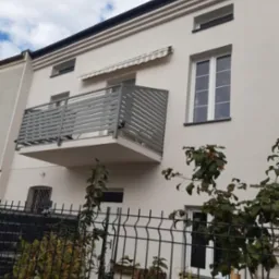 Elewacja jasnego budynku z szarym balkonem z poziomymi panelami, widoczna markiza nad balkonem, okna i czarne wykończenie dachu, ogrodzenie panelowe na pierwszym planie.