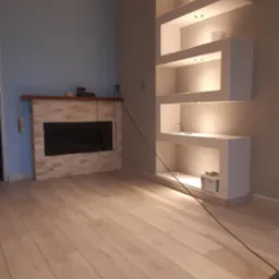 Nowoczesne wnętrze z kominkiem obłożonym kamieniem dekoracyjnym i wnęką ścienną z podświetlanymi półkami, widoczny kabel od odkurzacza i resztki po remoncie.