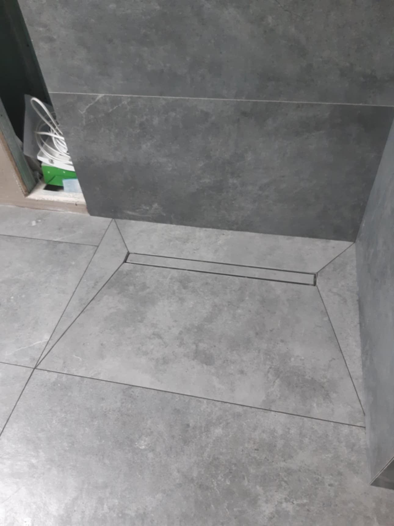 Szare płytki podłogowe w łazience z odpływem liniowym w rogu, widoczne przewody i puszka elektryczna.