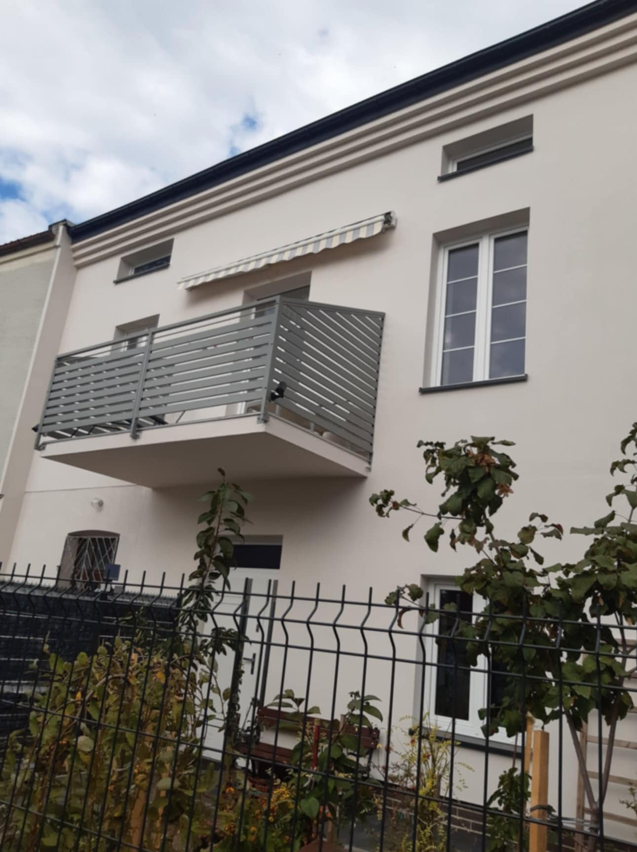 Elewacja jasnego budynku z szarym balkonem z poziomymi panelami, widoczna markiza nad balkonem, okna i czarne wykończenie dachu, ogrodzenie panelowe na pierwszym planie.