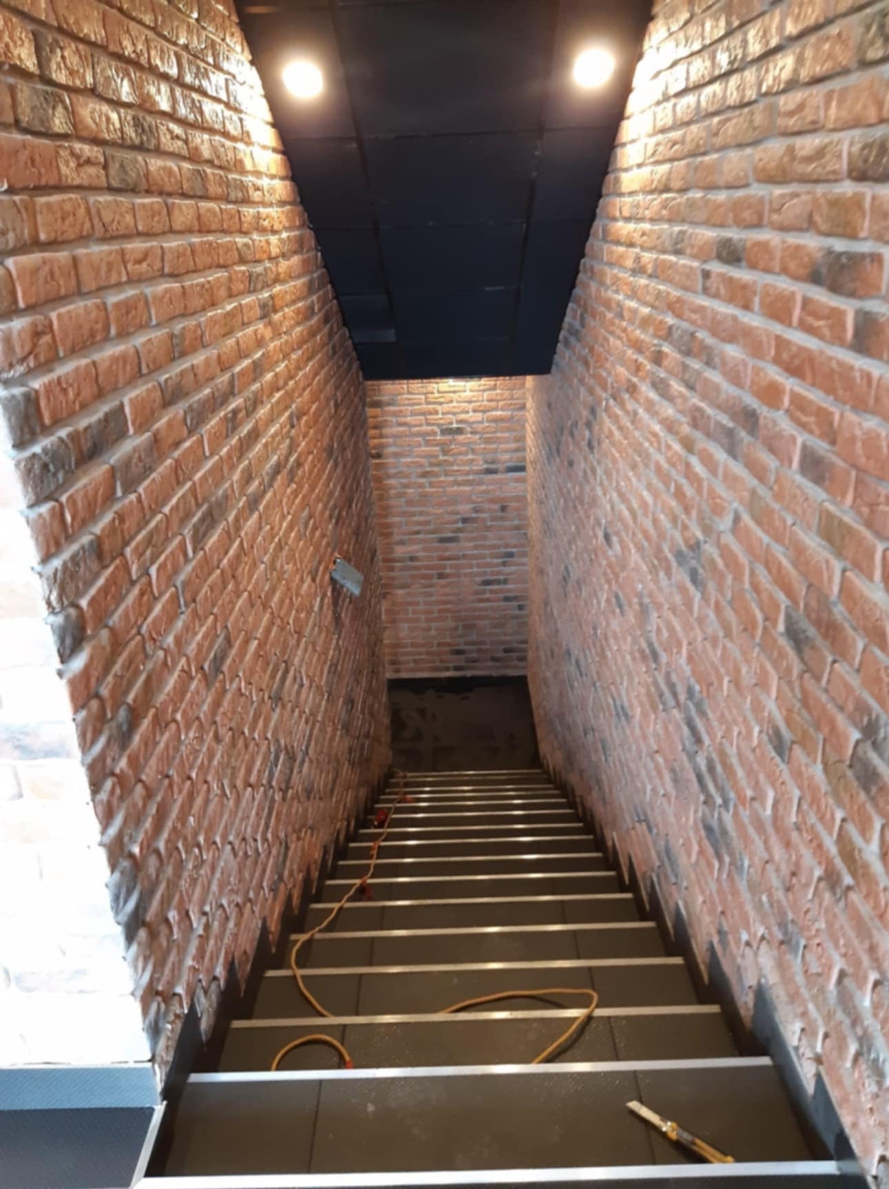 Klatka schodowa wykończona cegłą klinkierową w odcieniach czerwieni i brązu, prowadząca w dół, oświetlona punktowymi lampami sufitowymi, z widocznym kablem i narzędziem na schodach.
