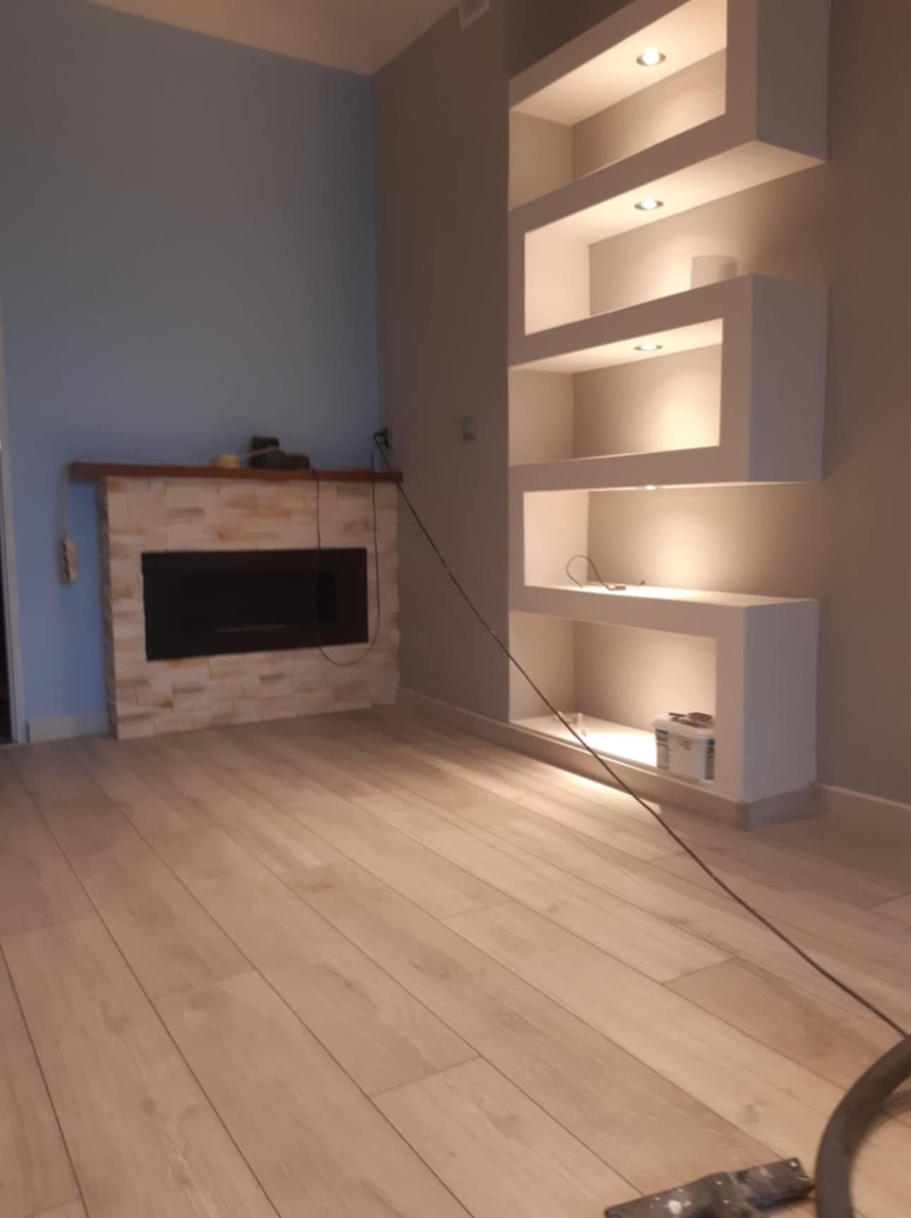 Nowoczesne wnętrze z kominkiem obłożonym kamieniem dekoracyjnym i wnęką ścienną z podświetlanymi półkami, widoczny kabel od odkurzacza i resztki po remoncie.