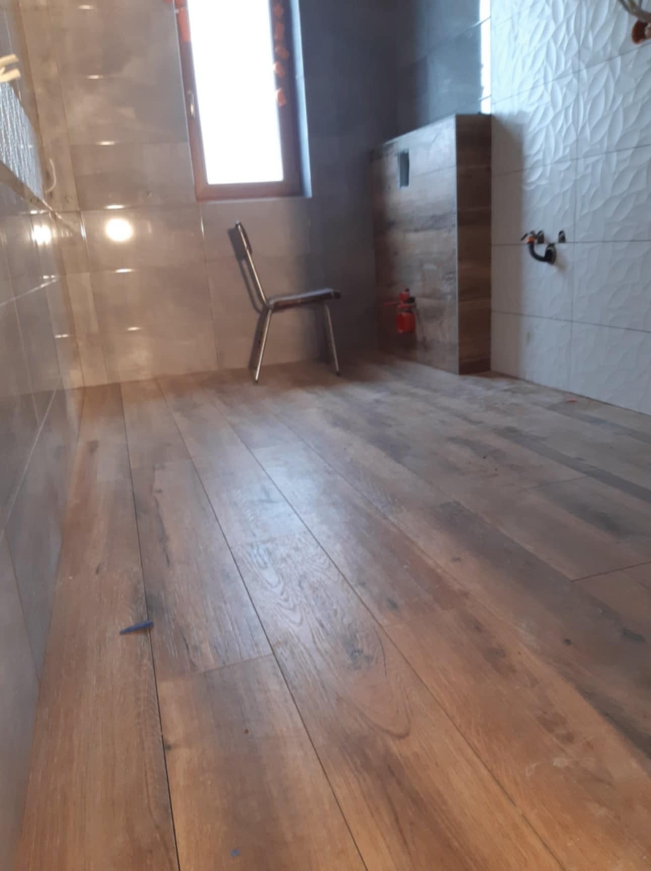 Wnętrze łazienki w trakcie wykańczania: drewnopodobne płytki podłogowe, częściowo wykończona zabudowa toalety, krzesło przy oknie.