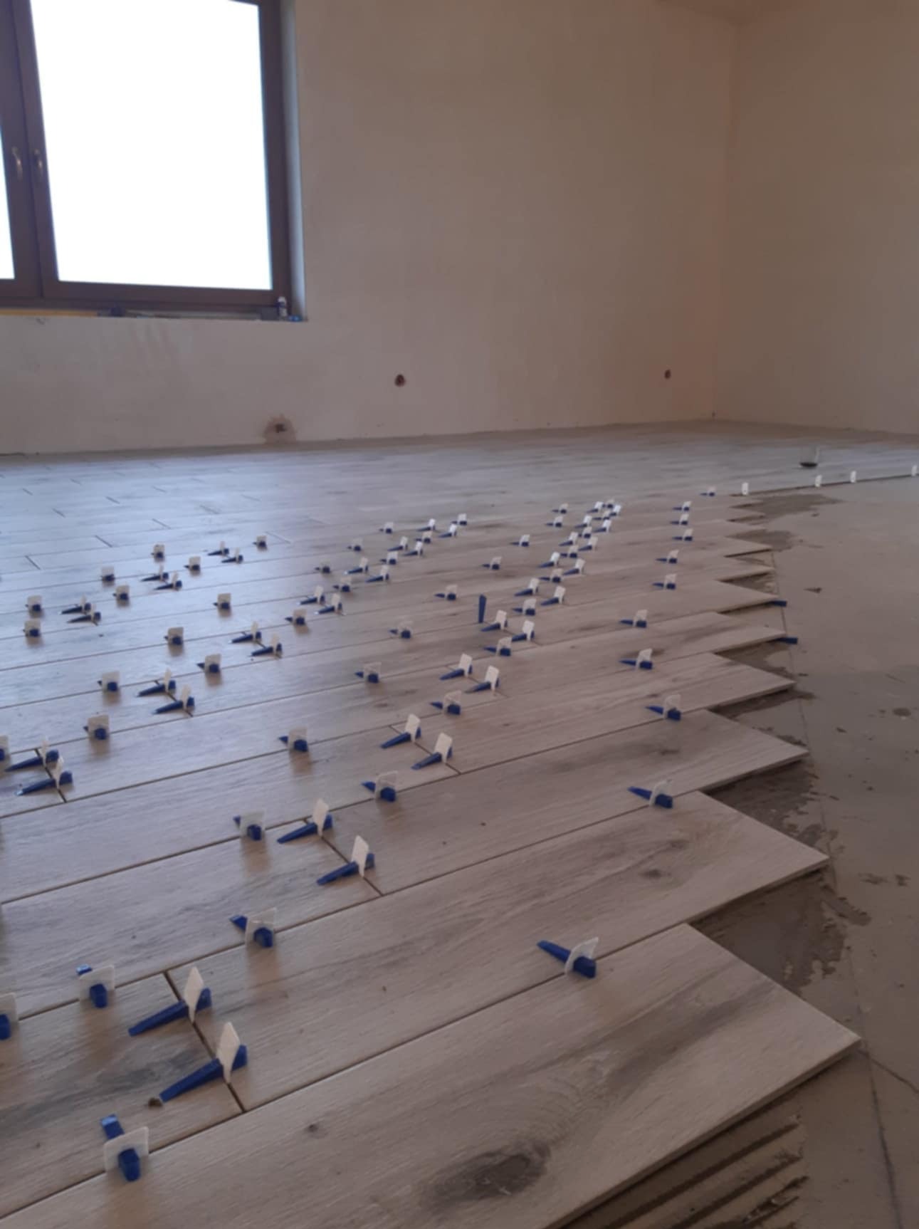 Układanie podłogi z płytek imitujących drewno, z użyciem niebieskich klinów poziomujących, widok na częściowo wykończoną powierzchnię i okno w tle.