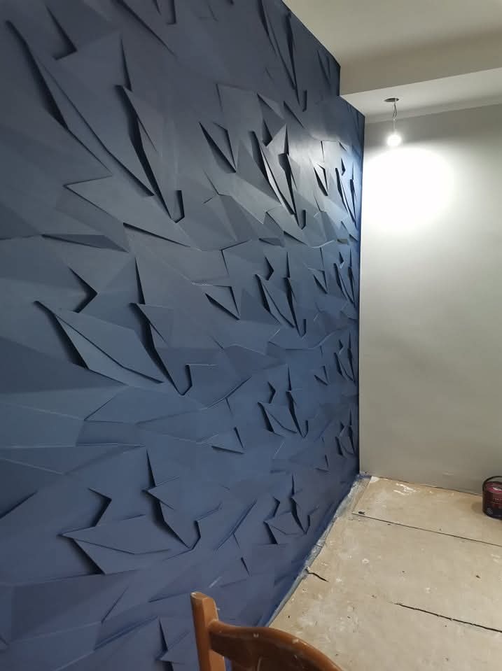 Niebieska ściana z trójwymiarowymi panelami dekoracyjnymi o geometrycznym wzorze w nowym pomieszczeniu z gołą żarówką i widoczną podłogą w trakcie wykończenia.