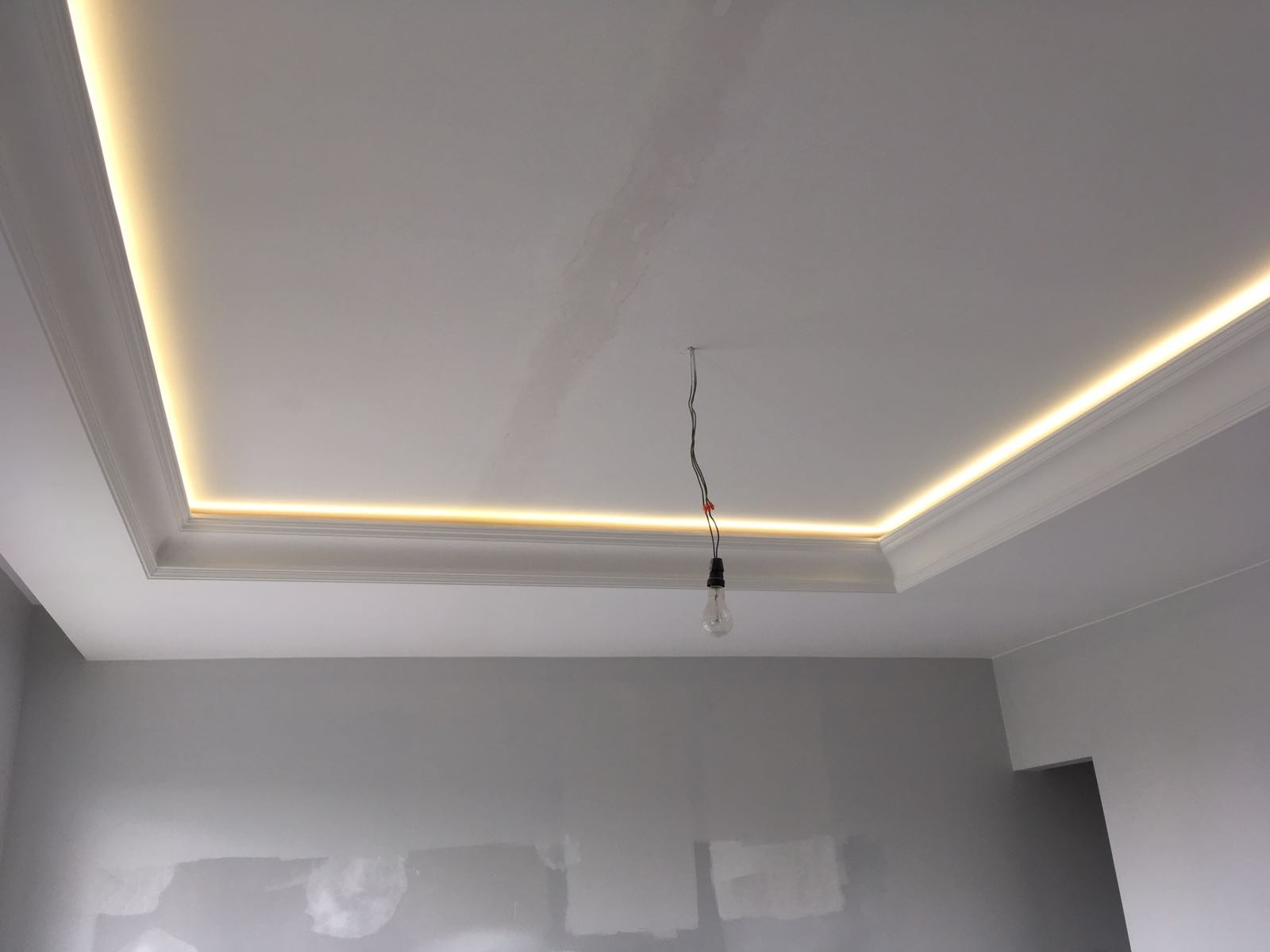 Szara ściana w trakcie malowania, sufit z listwami dekoracyjnymi i oświetleniem LED, widoczna wisząca żarówka tymczasowa.