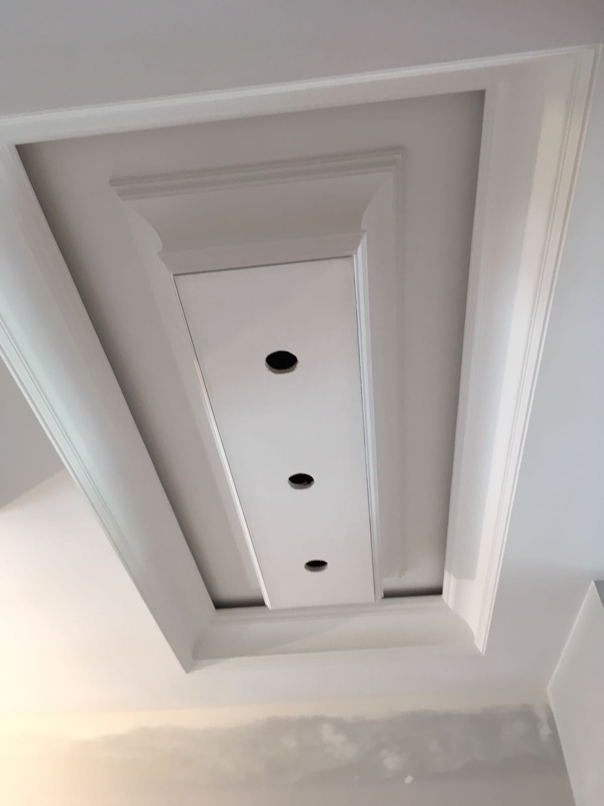 Kaseton sufitowy z trzema otworami na oświetlenie punktowe, ozdobiony sztukaterią w kolorze białym, widok z dołu na tle ściany w trakcie prac wykończeniowych.