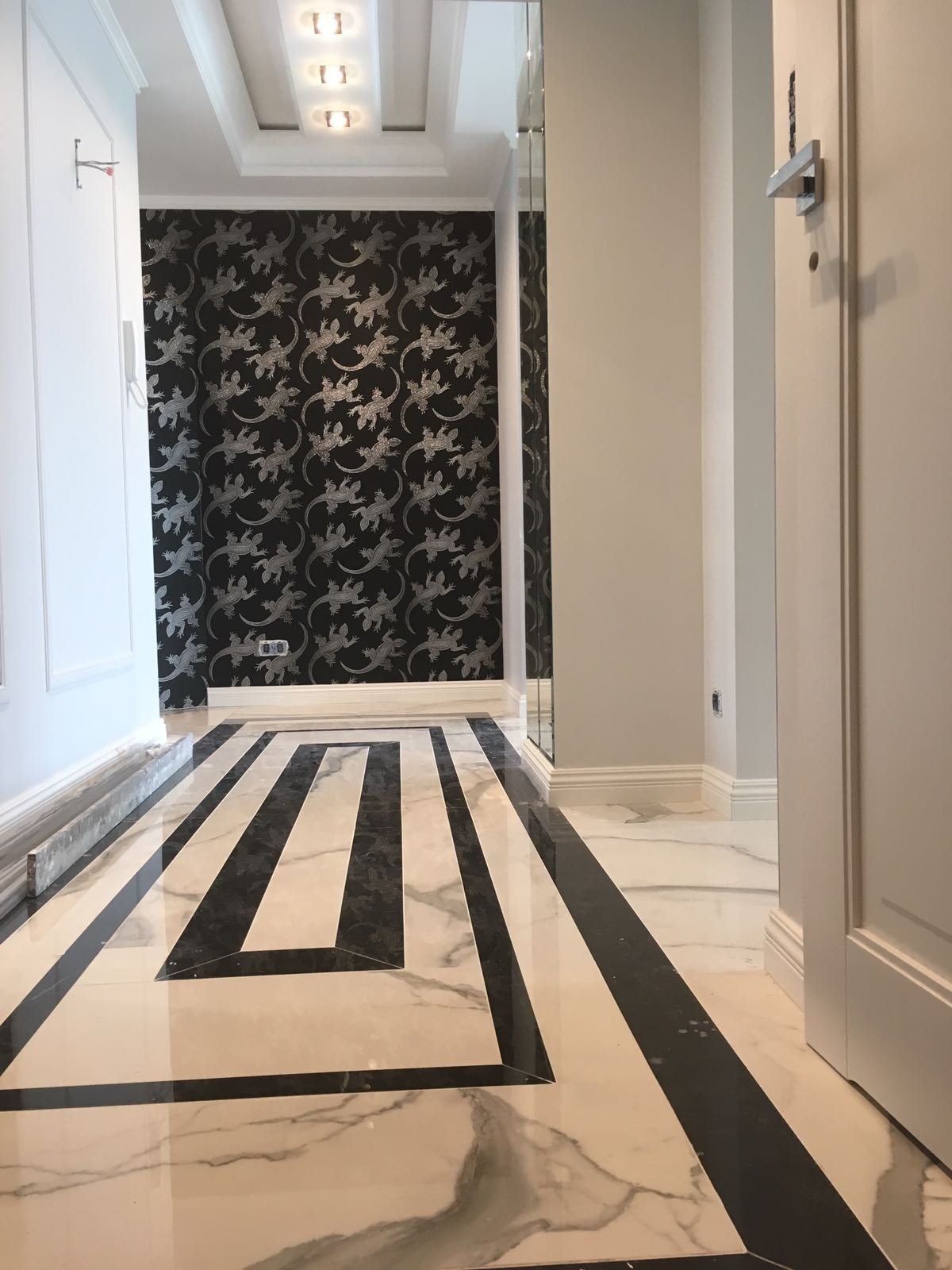 Wnętrze korytarza z marmurową podłogą w geometryczne wzory, ściana w czarno-srebrną tapetę z motywem jaszczurek, oświetlenie sufitowe punktowe.