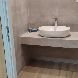 Usługi Remontowe Paweł Czerwiński - Wykończona łazienka z umywalką nablatową, szarą płytką na ścianach i drewnopodobną na podłodze. Widoczne kable wystające ze ściany, sugerujące przyszłe oświetlenie.