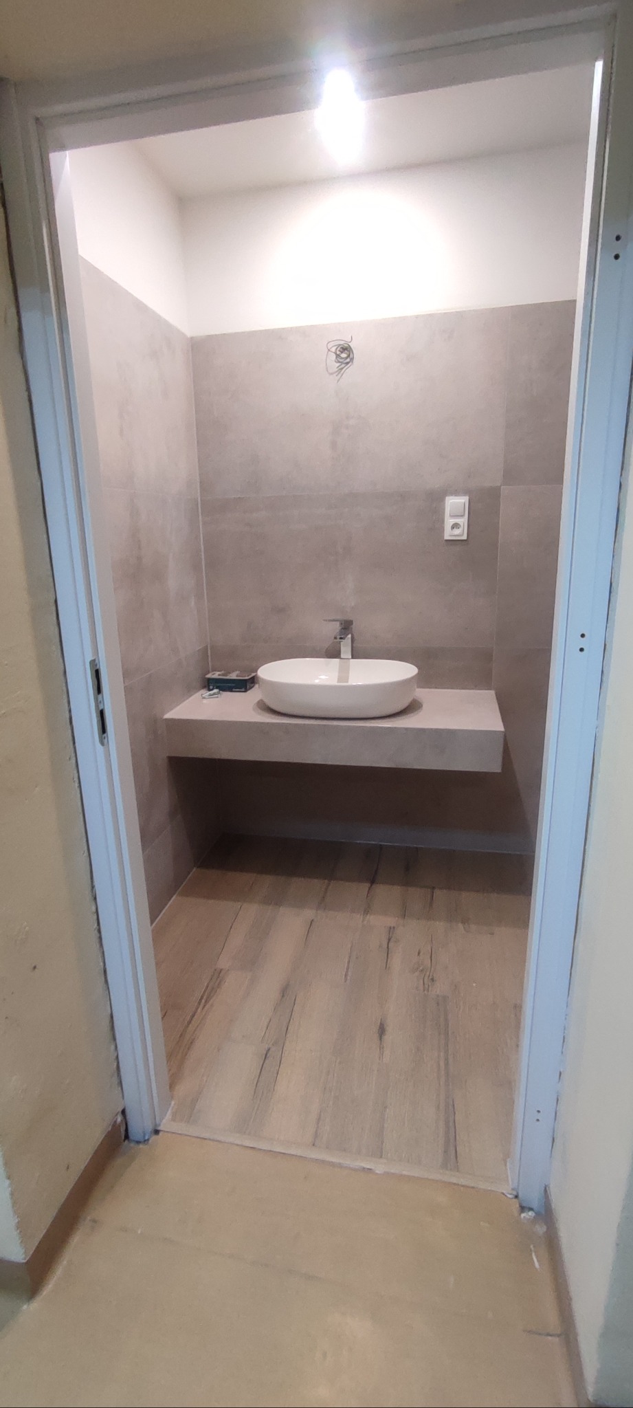 Wykończona łazienka z umywalką nablatową na blacie z płytek, drewnopodobna podłoga, szare płytki na ścianach. Widoczne przewody elektryczne do oświetlenia.