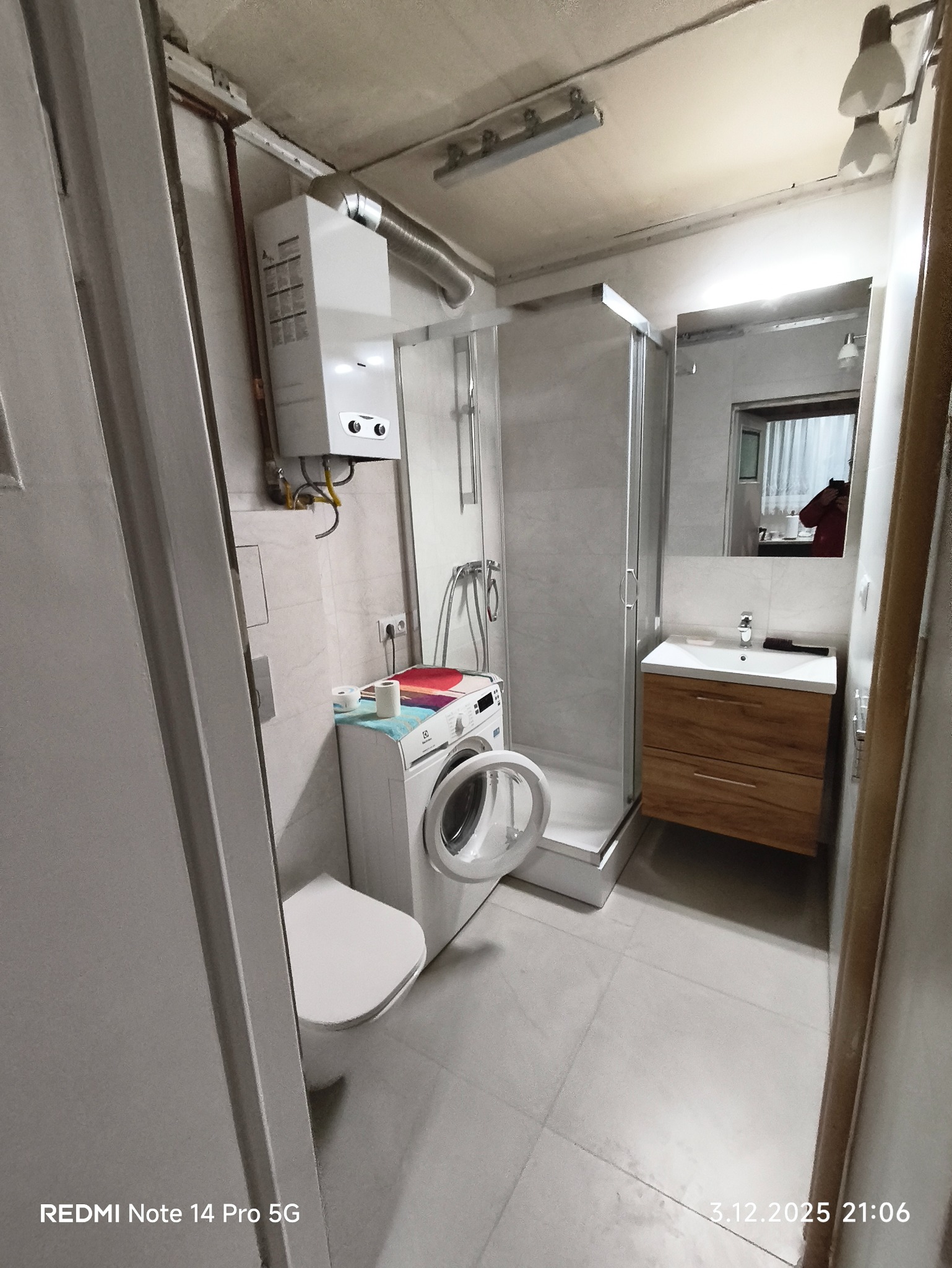 Wykończona łazienka z kabiną prysznicową, umywalką z szafką, toaletą i pralką. Na ścianie zamontowany bojler. Minimalistyczny styl, jasne płytki podłogowe.