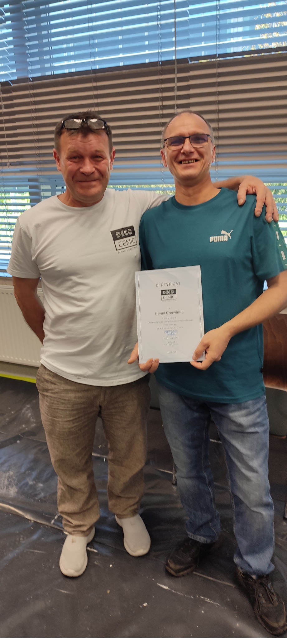 Dwóch uśmiechniętych mężczyzn pozuje z certyfikatem DECO CEMIC na tle okna z żaluzjami. Jeden z nich ma ramię na ramieniu drugiego.