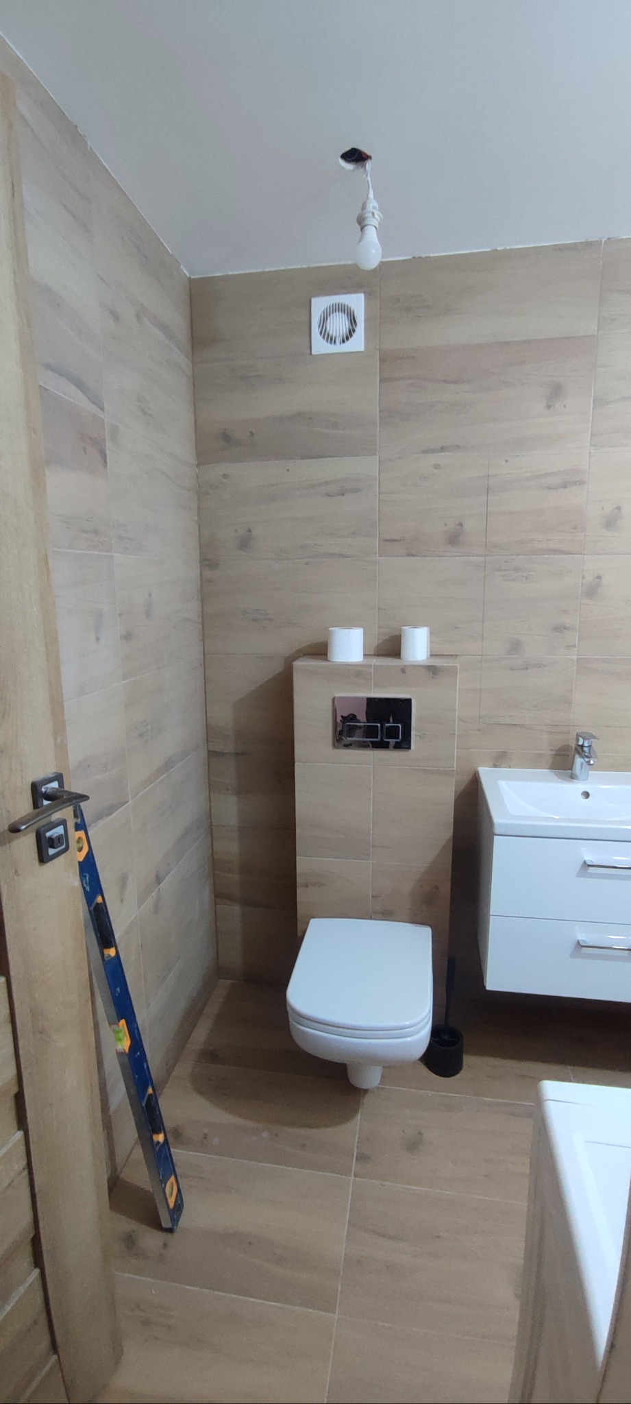 Wykończona łazienka z drewnopodobnymi płytkami, białą armaturą i poziomnicą opartą o futrynę. Widoczny wentylator i żarówka bez klosza.