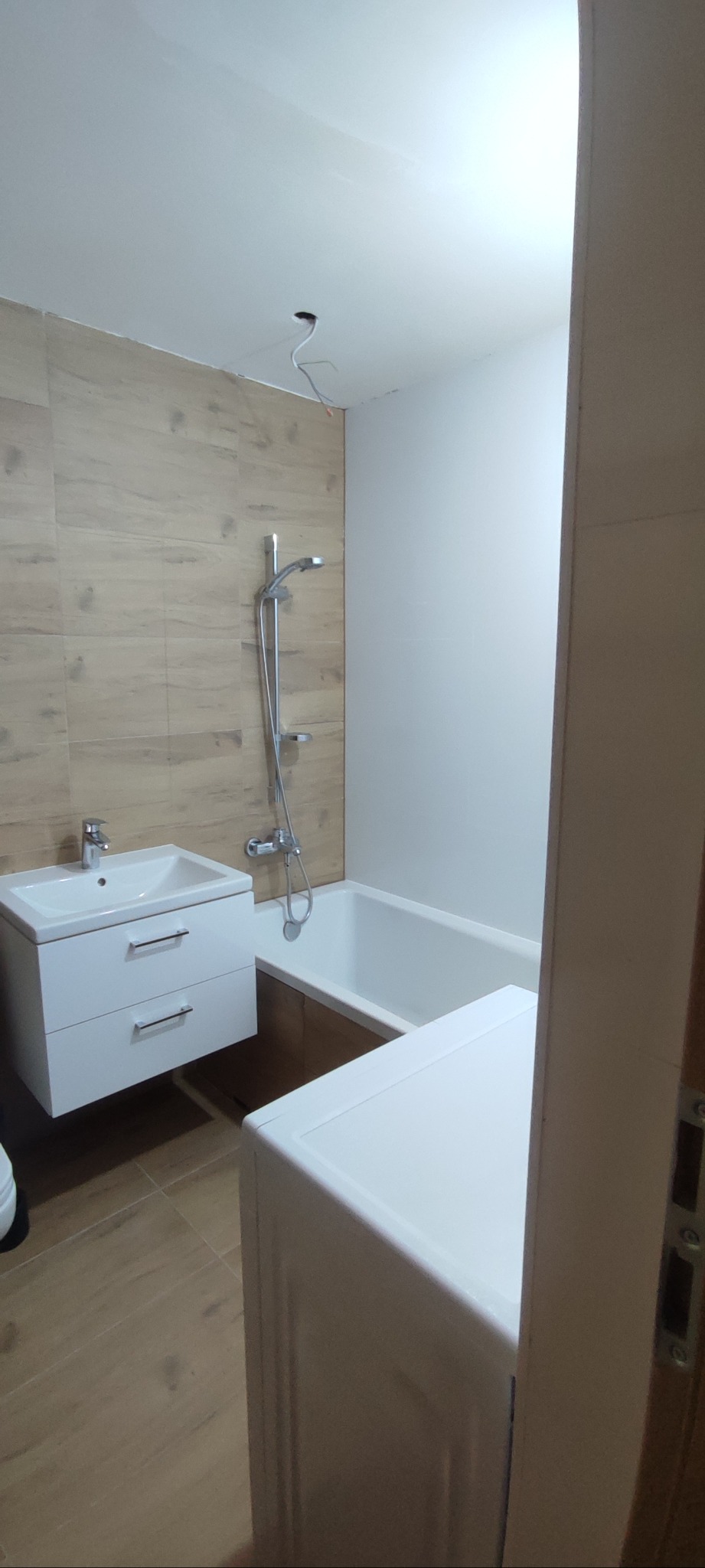 Wykończona łazienka z drewnopodobnymi płytkami, białą armaturą i pralką. Widoczne podłączenie elektryczne w suficie. Ujęcie z perspektywy wejścia.