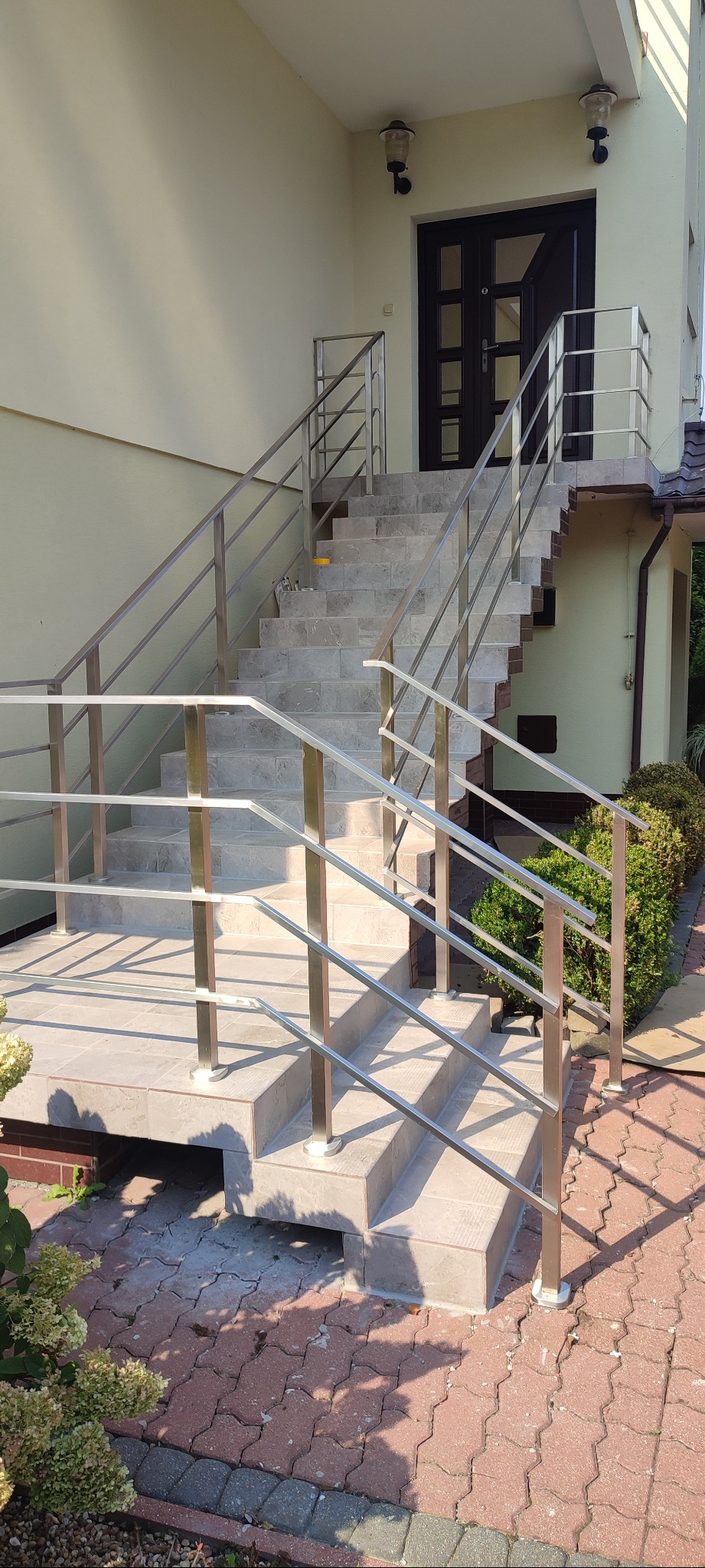 Zewnętrzne schody z szarych płytek prowadzące do ciemnych drzwi wejściowych, zabezpieczone nowoczesną, metalową balustradą. Widoczne lampy zewnętrzne i fragment elewacji budynku.