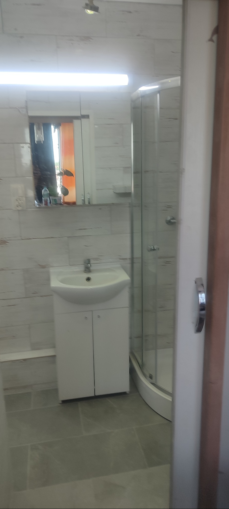 Jasna łazienka po remoncie: białe płytki imitujące drewno, kabina prysznicowa z zaokrąglonym szkłem, umywalka z szafką. Widoczne lustro i oświetlenie.