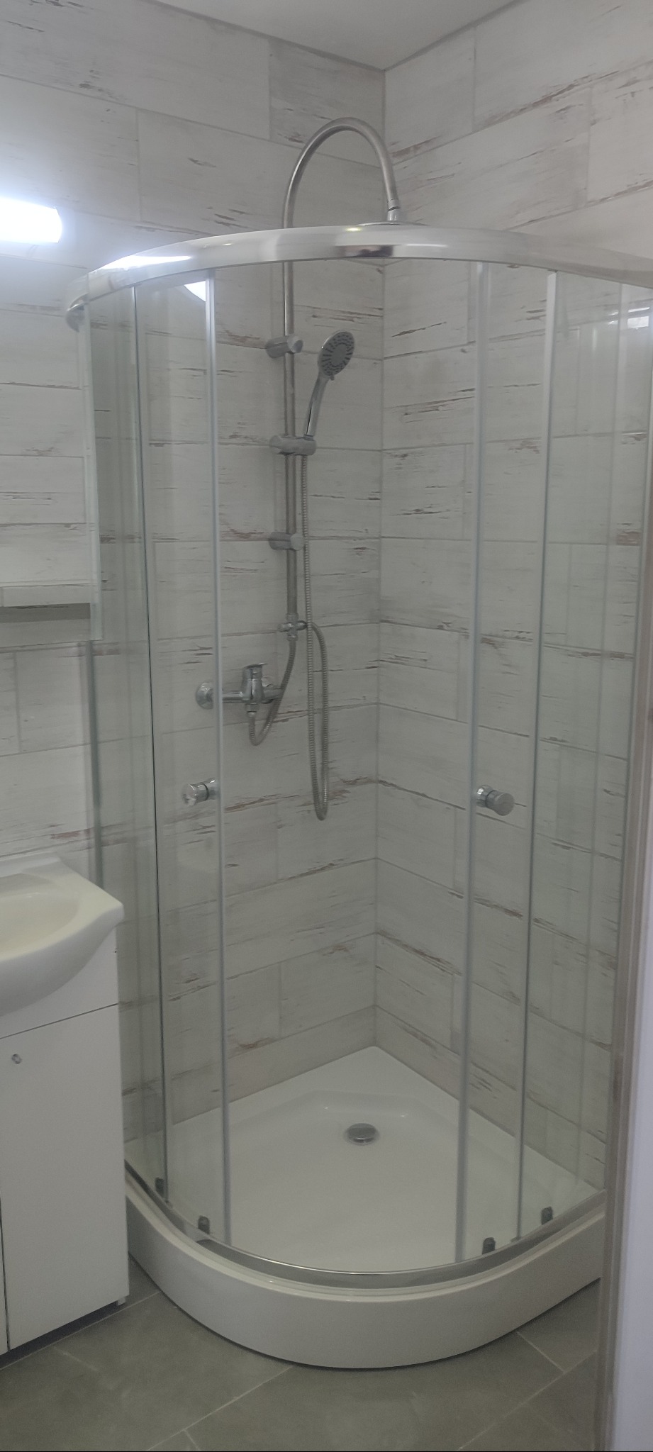 Narożna kabina prysznicowa z chromowanymi profilami i deszczownicą, na tle ściany wyłożonej płytkami imitującymi drewno. Widoczna umywalka z szafką.