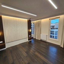 EKONEX SPÓŁKA Z OGRANICZONĄ ODPOWIEDZIALNOŚCIĄ - Nowoczesna sypialnia z ciemną drewnianą podłogą, panelem tapicerowanym za łóżkiem z podświetleniem LED, wiszącymi szafkami nocnymi i beżowymi zasłonami w oknach.