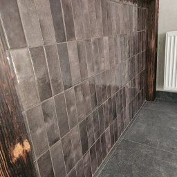 EKONEX SPÓŁKA Z OGRANICZONĄ ODPOWIEDZIALNOŚCIĄ - Ściana wyłożona pionowymi, szarymi płytkami z ciemnobrązową, drewnianą ramą; szara podłoga z widocznym grzejnikiem w tle.