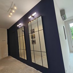 EKONEX SPÓŁKA Z OGRANICZONĄ ODPOWIEDZIALNOŚCIĄ - Korytarz w trakcie remontu z ciemnogranatową ścianą, dwoma wysokimi lustrami z czarnymi szprosami, surową podłogą i fragmentem ściany z widocznymi śladami po gładzi.