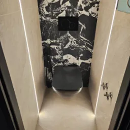 Nowoczesna toaleta z czarną miską wiszącą i spłuczką podtynkową na tle ściany wyłożonej czarno-białym marmurem, oświetlona ledowymi listwami wzdłuż beżowych ścian.