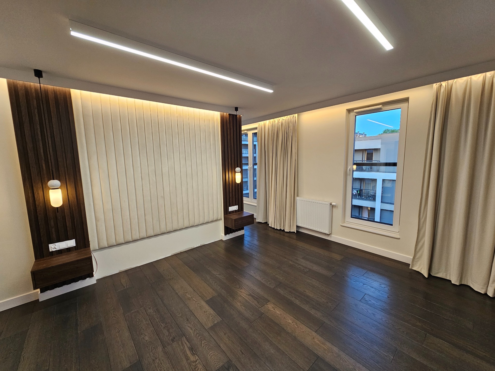 Nowoczesna sypialnia z ciemną drewnianą podłogą, panelem tapicerowanym za łóżkiem z podświetleniem LED, wiszącymi szafkami nocnymi i beżowymi zasłonami w oknach.