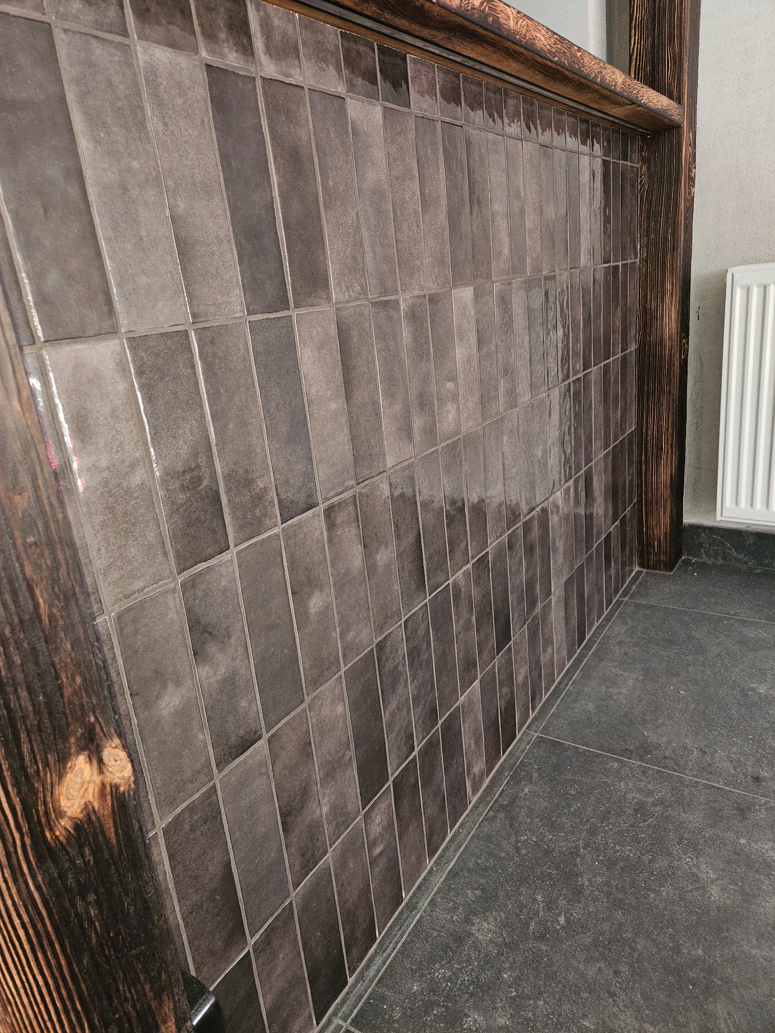 Ściana wyłożona pionowymi, szarymi płytkami z ciemnobrązową, drewnianą ramą; szara podłoga z widocznym grzejnikiem w tle.