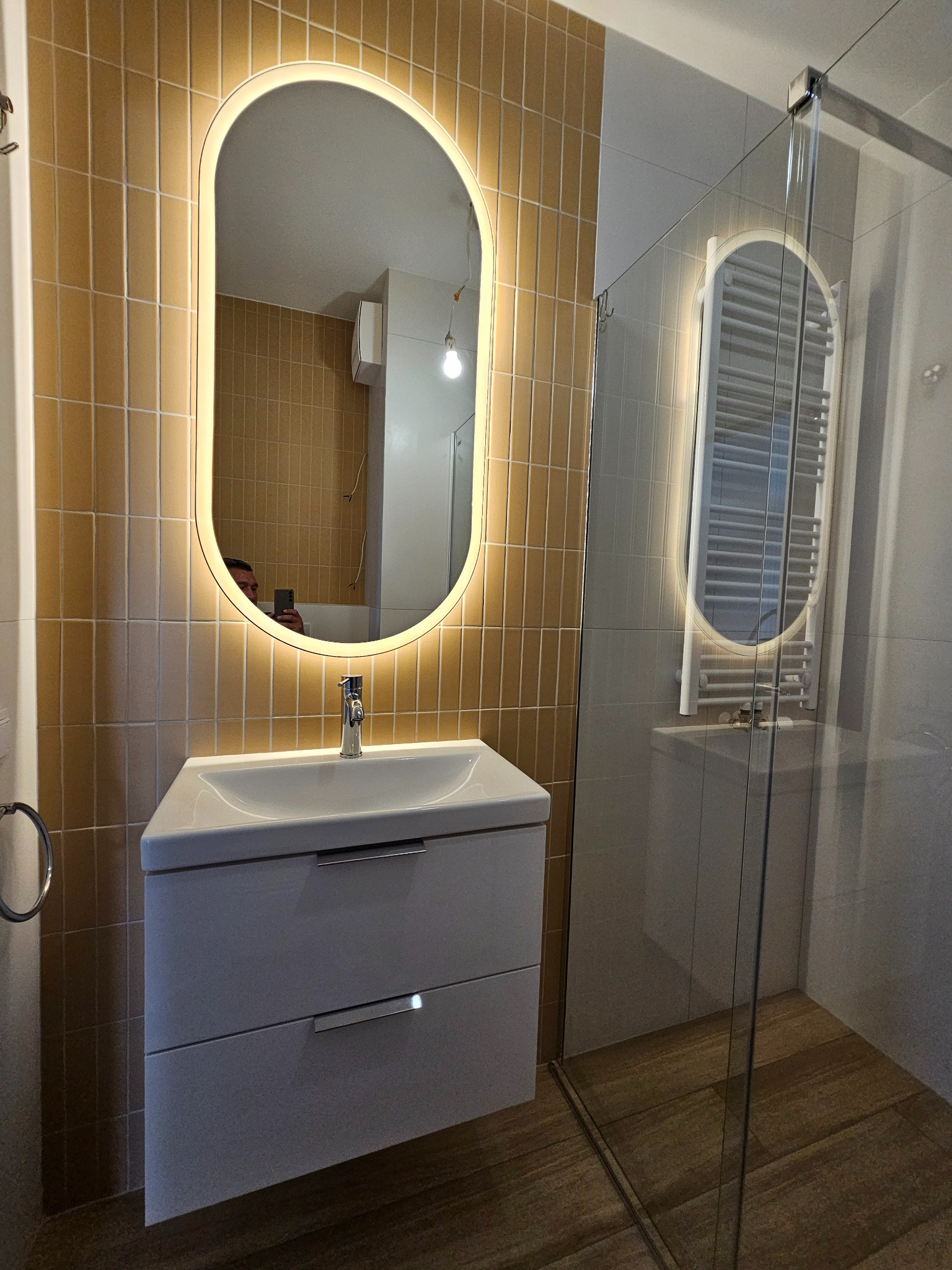 Nowoczesna łazienka w Krakowie z beżowymi, pionowo ułożonymi płytkami, podświetlanym owalnym lustrem nad białą szafką z umywalką oraz kabiną prysznicową ze szklanymi drzwiami.