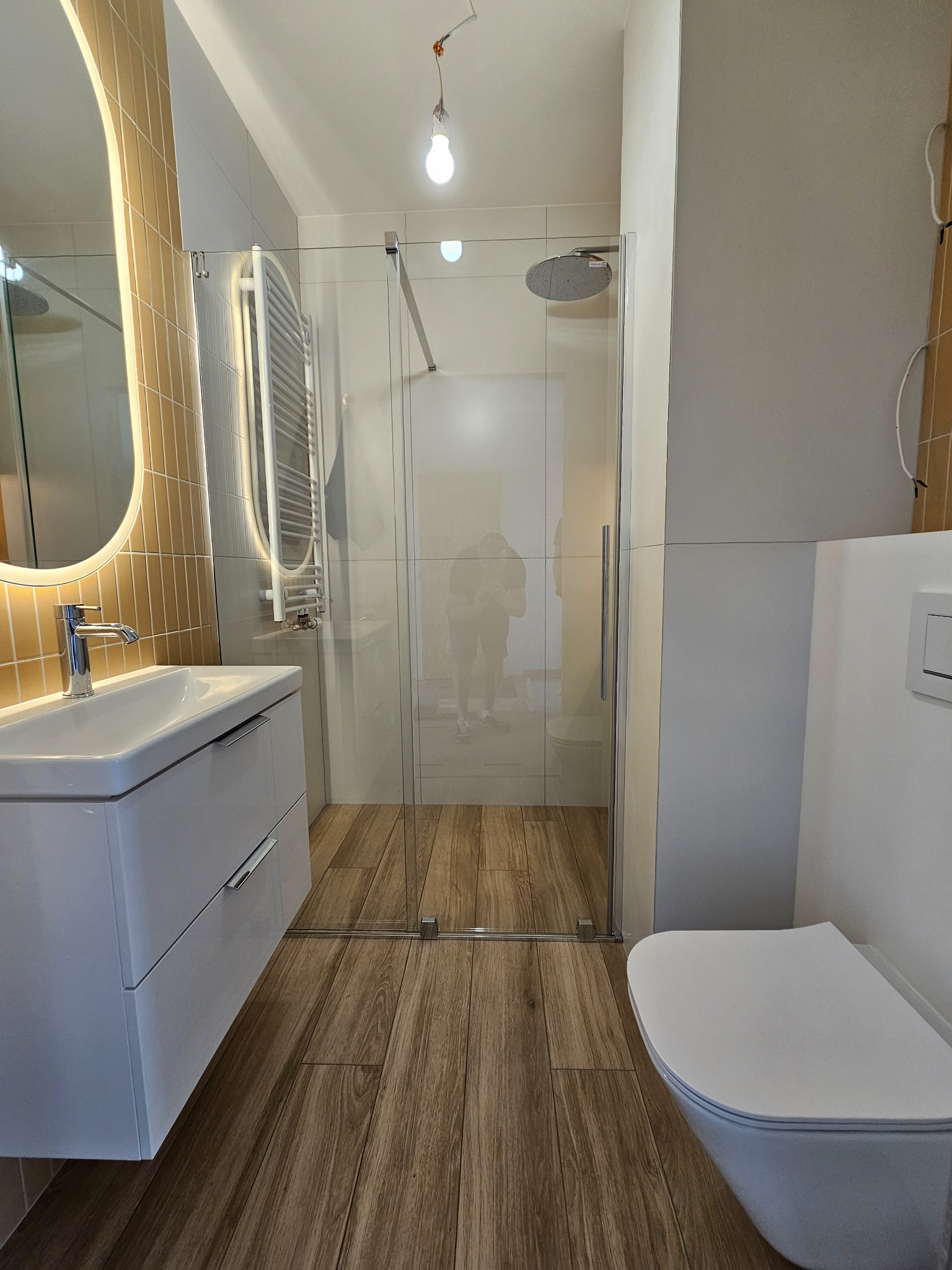 Nowoczesna łazienka z kabiną prysznicową typu walk-in, umywalką z szafką w kolorze białym, drewnopodobną podłogą i okrągłym lustrem z podświetleniem LED na mozaikowej ścianie.