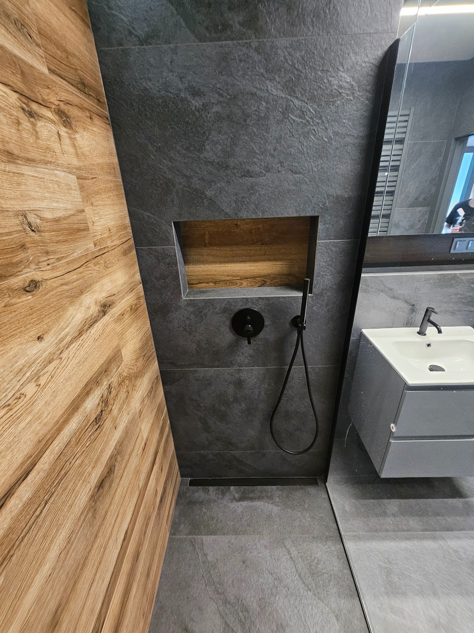Nowoczesna łazienka z drewnianą ścianą, szarą płytką imitującą kamień, czarną armaturą i wnęką z drewnianym wykończeniem.