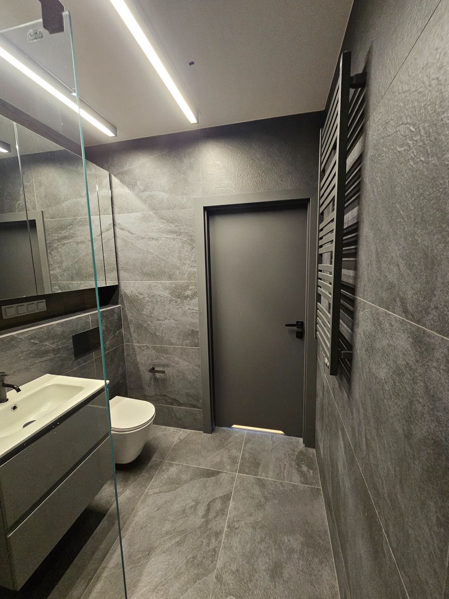 Nowoczesna łazienka w odcieniach szarości z płytkami imitującymi kamień, podświetlanym lustrem, grzejnikiem drabinkowym i minimalistyczną armaturą.