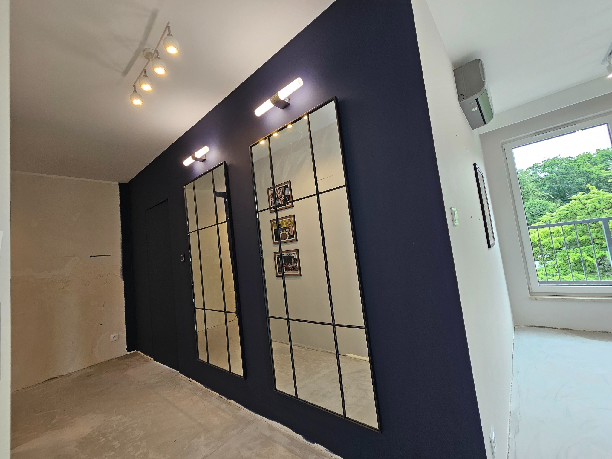 Korytarz w trakcie remontu z ciemnogranatową ścianą, dwoma wysokimi lustrami z czarnymi szprosami, surową podłogą i fragmentem ściany z widocznymi śladami po gładzi.
