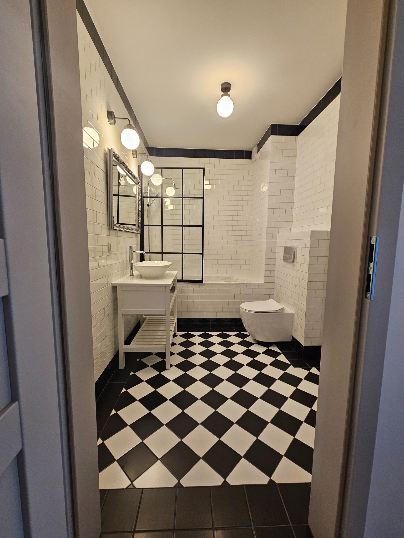 Nowoczesna łazienka z czarno-białą szachownicą na podłodze, białe płytki na ścianach i czarną ramą kabiny prysznicowej, widok zza uchylonych drzwi.