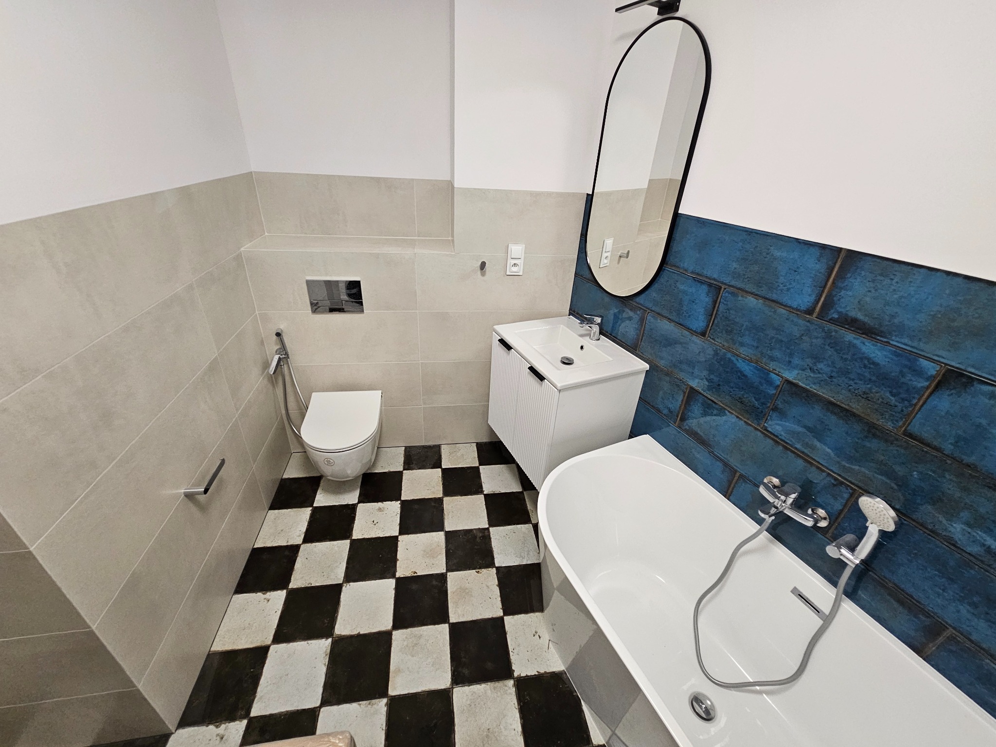 Wykończona łazienka z białą armaturą, umywalką z szafką, toaletą, wanną obudowaną niebieskimi płytkami imitującymi cegłę oraz podłogą w czarno-białą szachownicę.